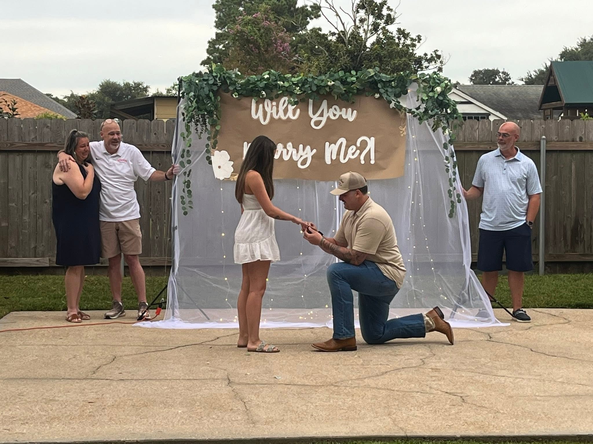 Shane David Hirstius drops to one knee to propose to his longtime love Leah Victoria Fonte during a backyard proposal setup. Wearing a beige collared shirt, jeans, boots, and a cap, Shane slips an engagement ring onto Leah’s finger as she stands before him in a white sundress in front of an arch adorned with greenery, fairy lights, and a banner reading "Will You Marry Me?!", while family members look on emotionally from either side. | Source: Facebook/StevenMarjorie Hirstius