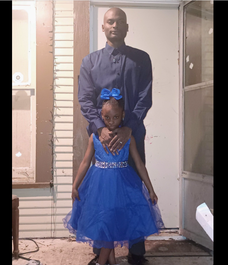 A photo of Shamar Elkins standing behind one of his daughters shows his hands gently resting near her shoulders as she faces the camera in a bright blue dress. | Source: Facebook/Trending News Tracker