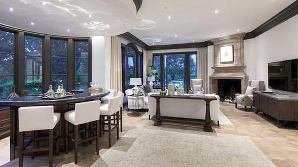 The estate's formal entertaining lounge features a dramatic curved dark-wood wet bar with cushioned barstools situated beneath a circular recessed ceiling, balanced across parquet wood flooring by a carved stone fireplace, cozy neutral seating, and tall arched French doors opening to the grounds. | Source: YouTube/Taste of Country