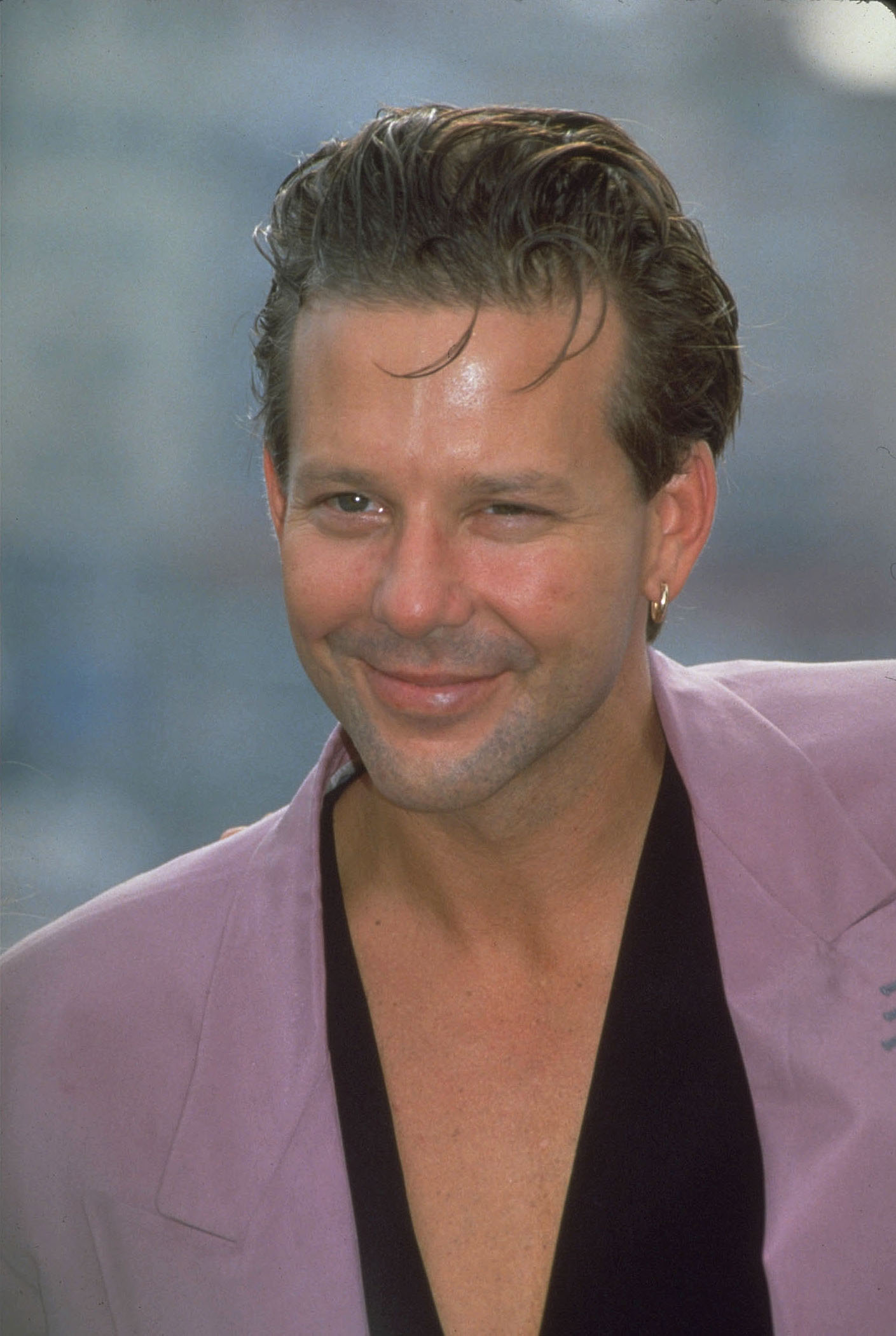 Mickey Rourke flashes a charismatic grin while exuding cool confidence in a lavender blazer and gold hoop earring. With tousled hair and a daring low-cut shirt, he perfectly embodies that era's Hollywood swagger.