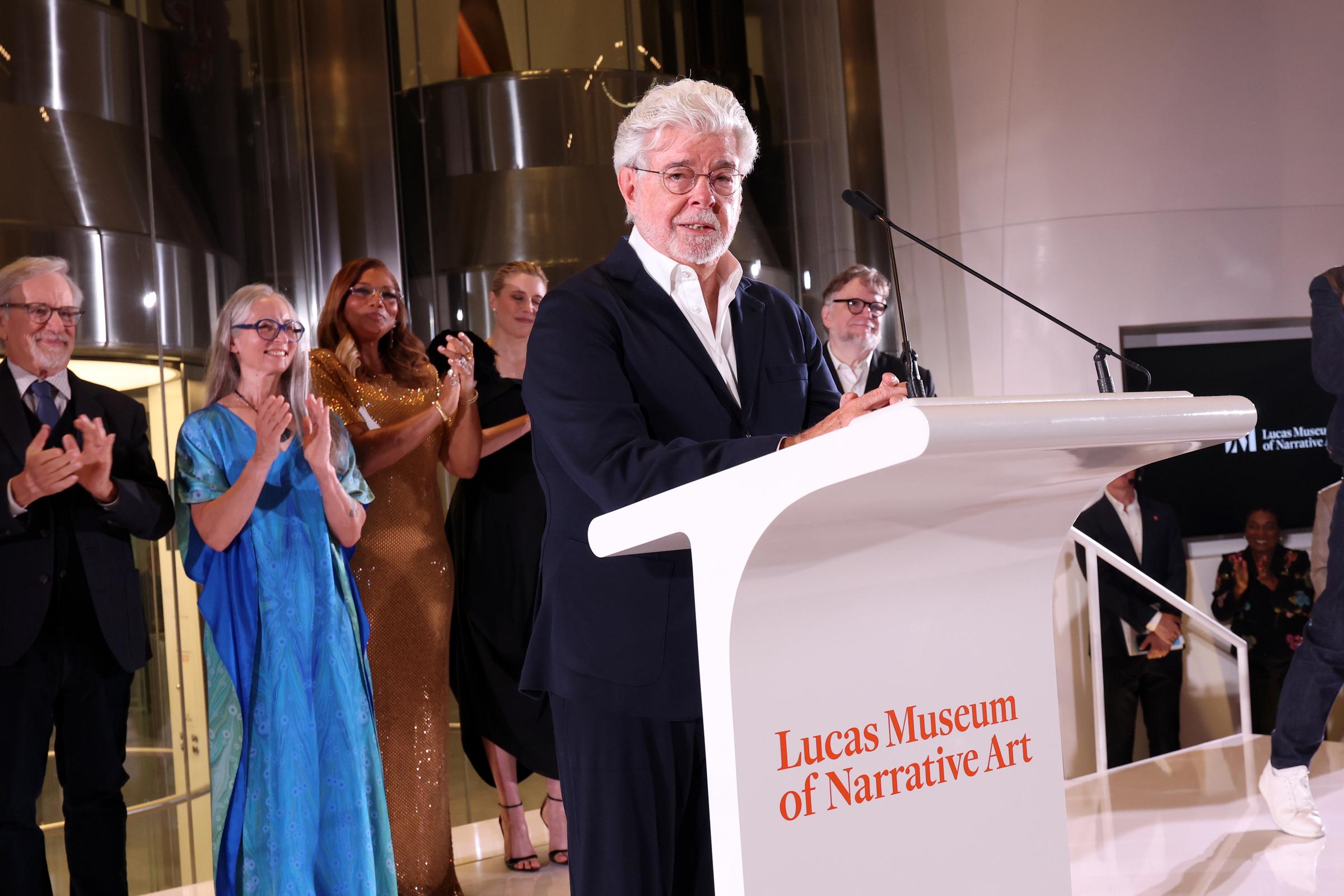 Lucas addressed guests from a podium bearing the Lucas Museum of Narrative Art name. Attendees behind him applauded during the opening-night program. | Source: Getty Images