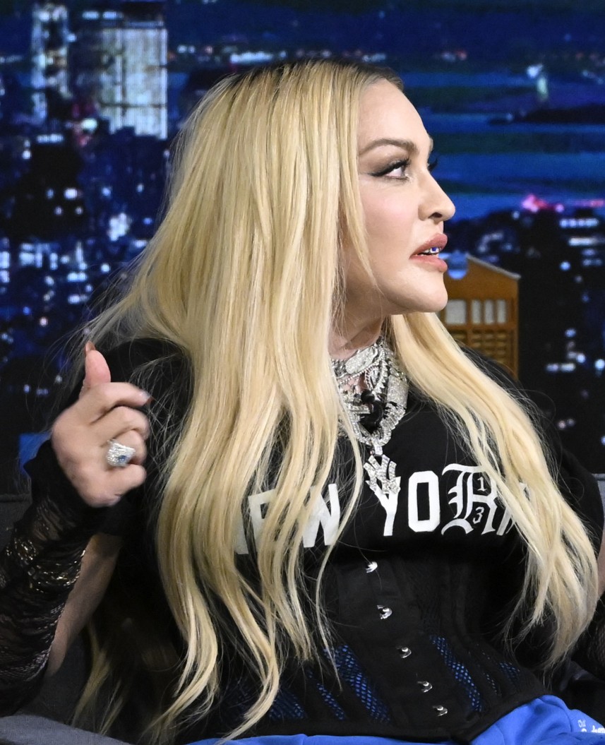 Madonna appears in profile during a television appearance with long blond hair, a black "New York" top, layered necklaces and a large statement ring. | Source: Chat GPT