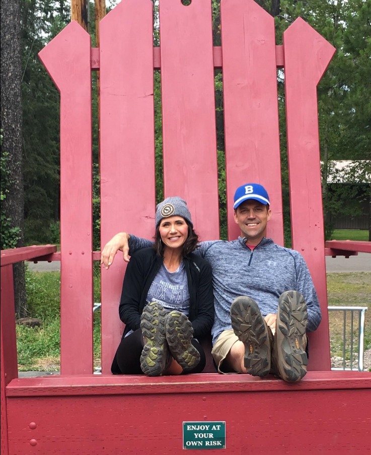 Relaxed and playful, Kristi Noem and her husband Bryon Noem lounge side by side on an oversized red chair, boots up and laughter easy. | Source: Facebook/KristiNoem