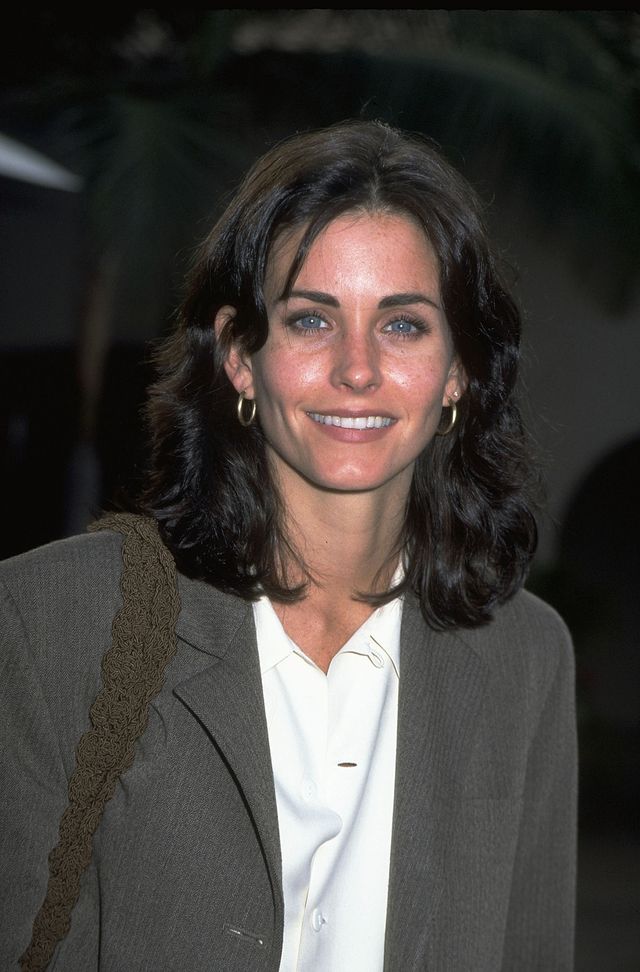 Courteney Cox wears light makeup, a crisp white shirt, and a gray coat to an event in 1999.