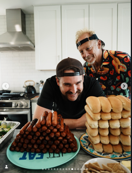 Two men pose behind a large stack of bread rolls and a platter piled with cooked sausages. White cabinets and stainless steel appliances form the backdrop, while the generous spread puts food at the center of the gathering. | Source: Instagram/wynonnajudd