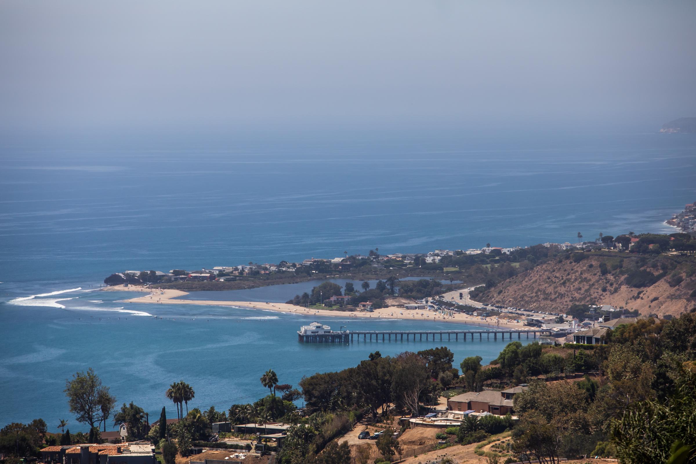 A scenic elevated view captured the historic Malibu Pier extending into the azure Pacific Ocean alongside the sandy curves of Malibu Lagoon State Beach on August 21, 2026, in Malibu, California. Gentle white surf curved along the coastline, with hillside greenery, coastal roadways, and beachfront properties stretching across the panoramic summer landscape. | Source: Getty Images