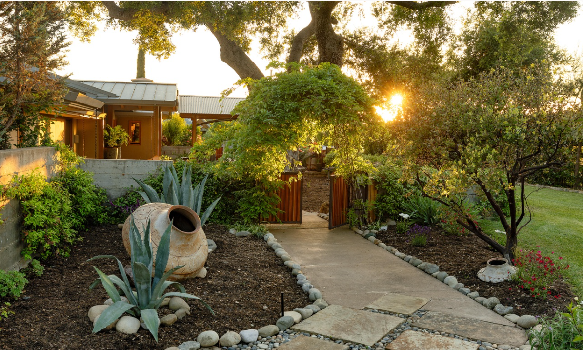 A stone pathway winds through a landscaped garden featuring agave plants, flowering shrubs, and mature trees beside the home. | Source: Redfin