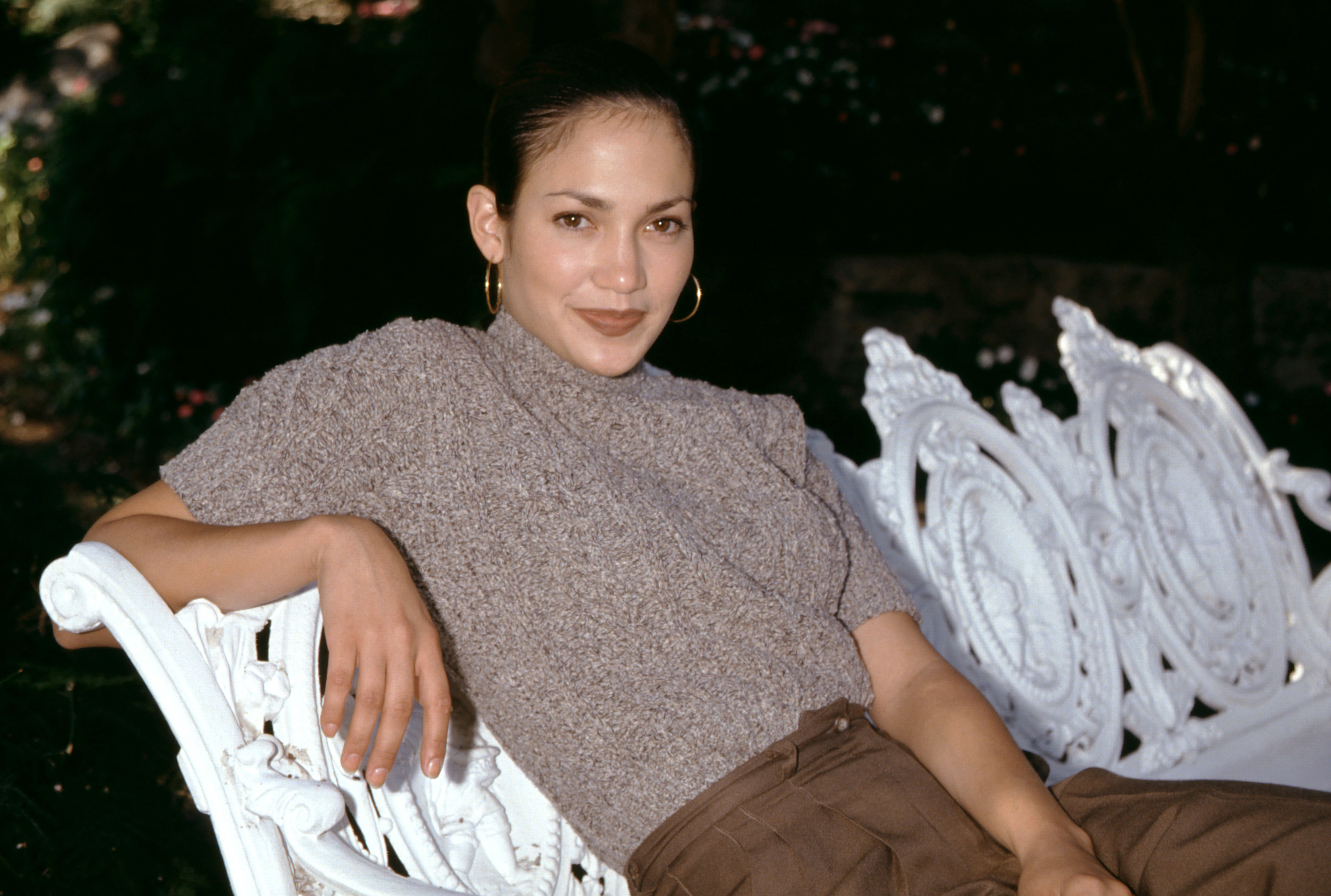 Jennifer Lopez reclines casually across an ornate white bench, resting one arm along the carved frame while wearing a textured high-neck top and loose brown trousers. With her slicked-back hair, hoop earrings, and calm expression, the portrait captures a relaxed yet polished early-career moment.