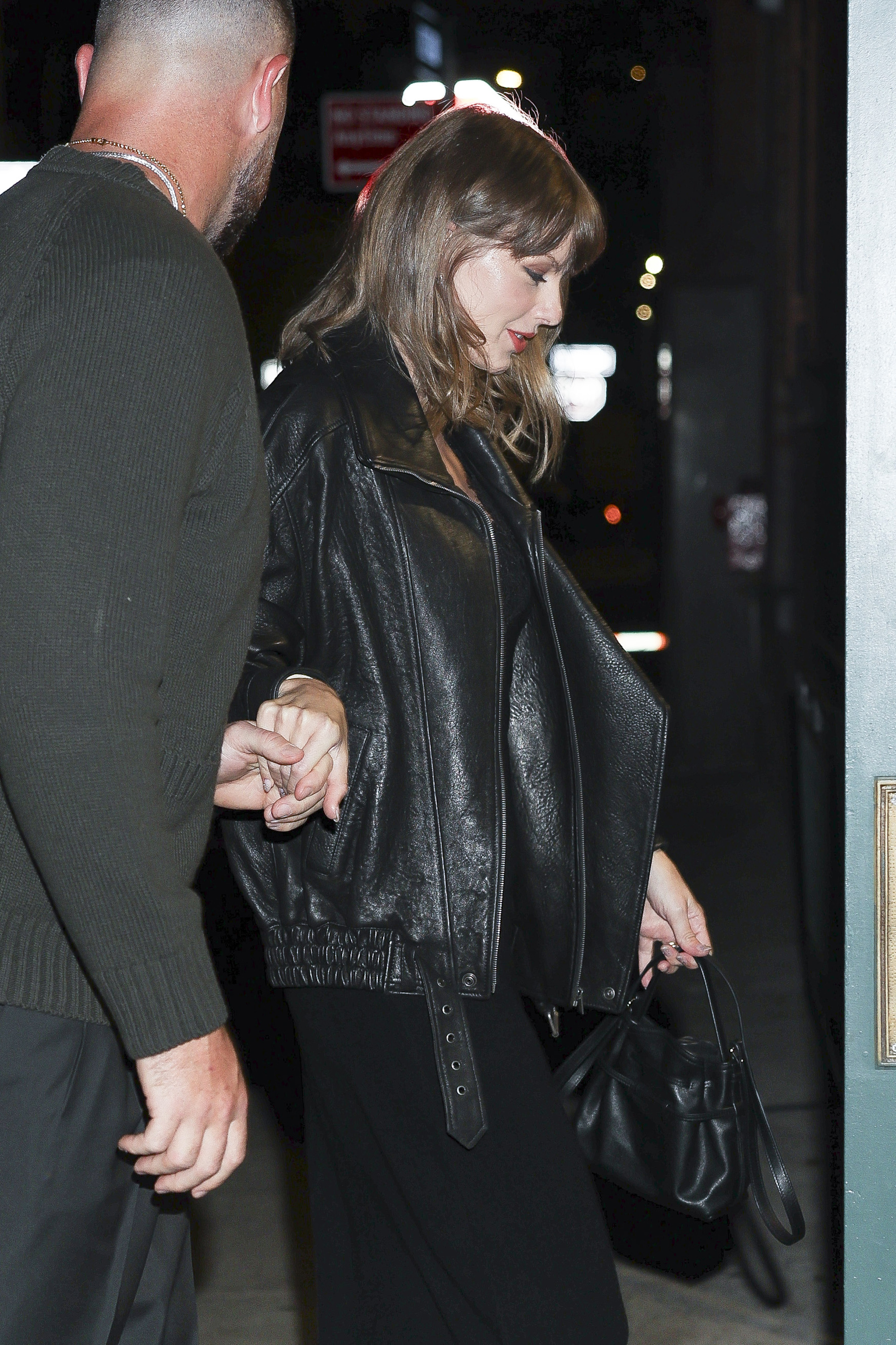 Swift holds Kelce's hand while carrying a black leather handbag, with her oversized leather jacket and signature red lipstick completing the look. | Source: Getty Images