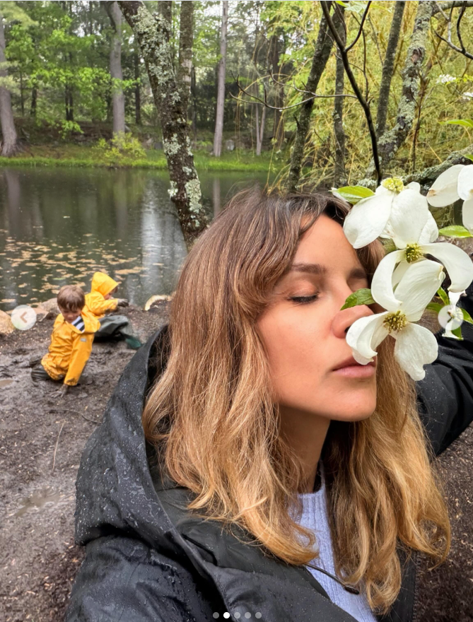 Alejandra Silva pauses beside a peaceful lakeside, holding a white flower close as her children play in the mud nearby. | Source: Instagram/alejandragere