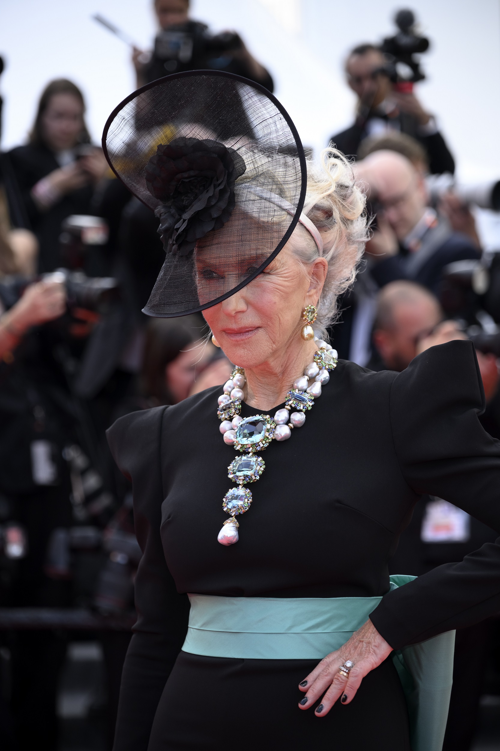 Mirren strikes a statuesque pose with one hand elegantly resting at her waist, fingers softly fanned to reveal a flash of rings and dark polish. Her stance exudes poise and control, lending regal authority to the drama of her hat and the luminous cascade of jewels at her neckline.