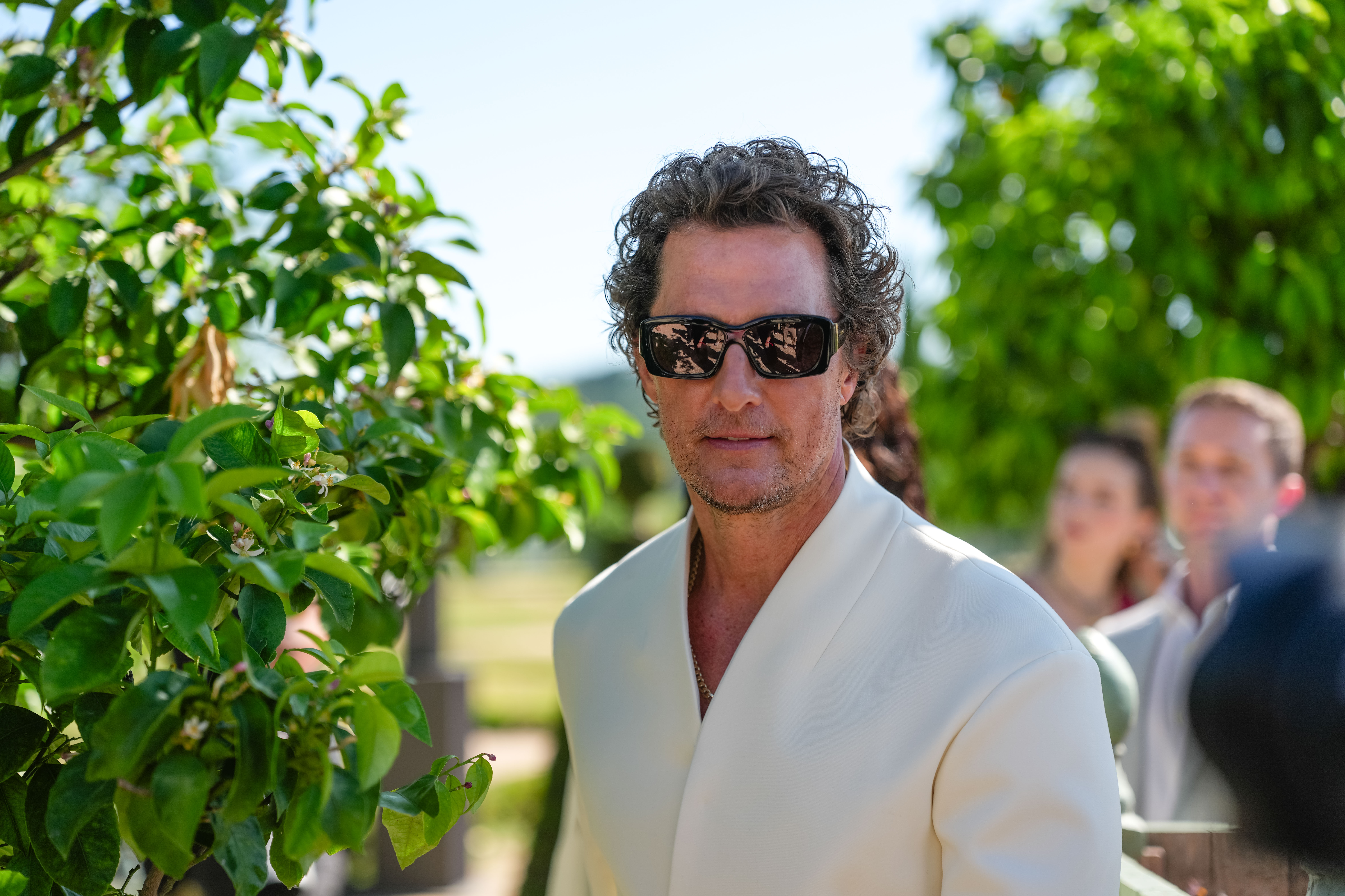 A close-up shot of McConaughey highlights the details of his accessories, with his black rectangular sunglasses featuring subtle gold Jacquemus branding on the temple. The photo also reveals a glimpse of a silver chain worn beneath his cream coat, adding a sleek, personal touch to his refined look.