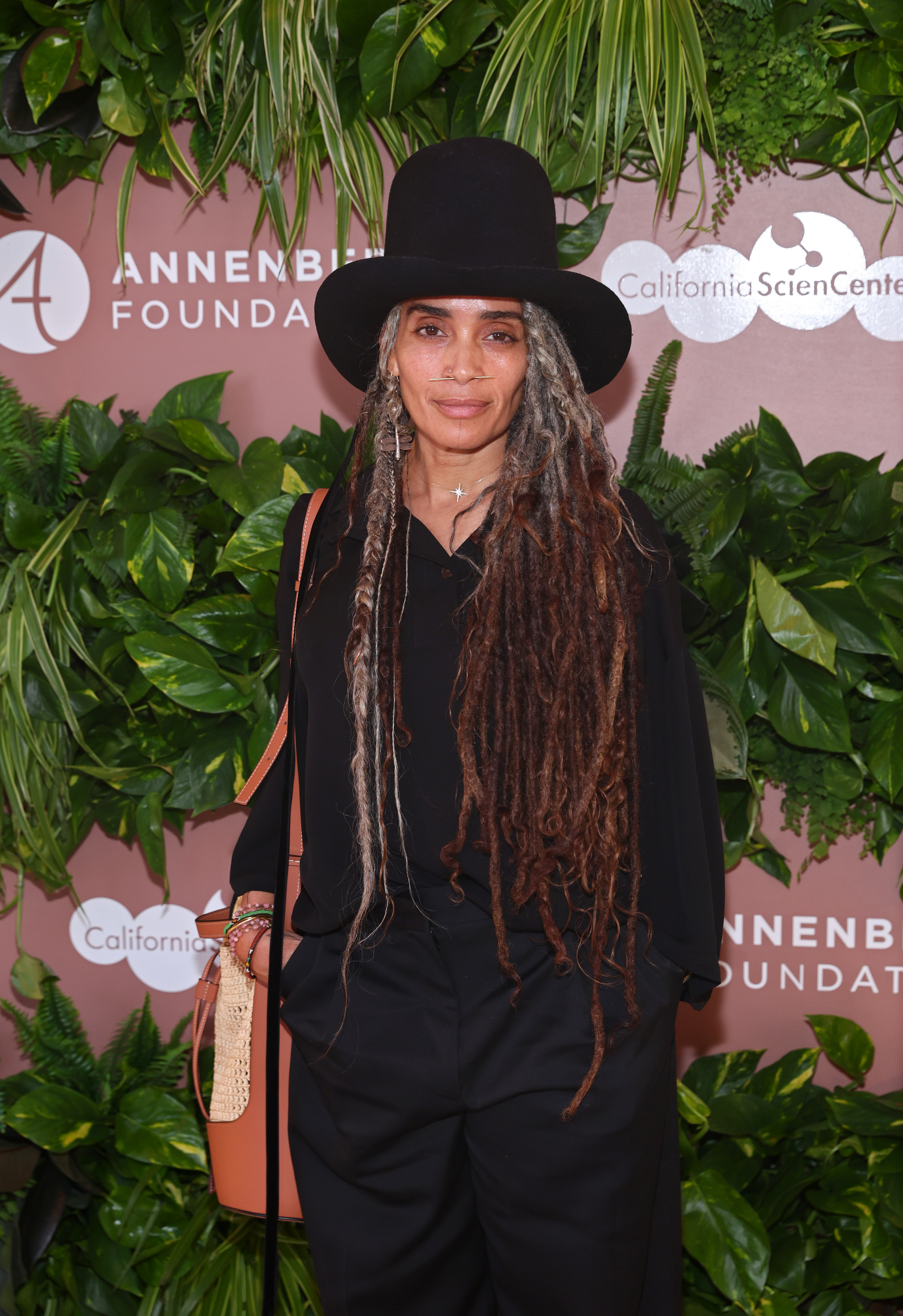 Lisa Bonet arrives at an Annenberg Foundation premiere event at the California Science Center, standing before a lush greenery backdrop in a relaxed all-black ensemble and wide-brim hat.